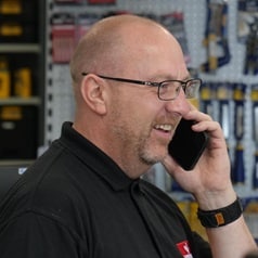 A Howdens worker stands in a depot underneath a big, red banner, taking a phone call.