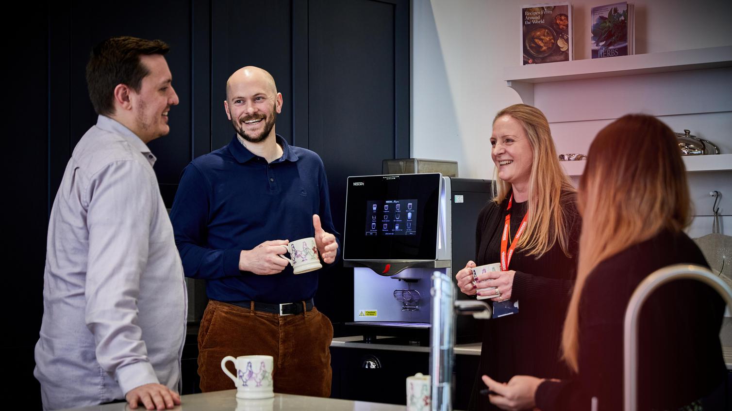 4 people chatting in the kitchen at Raunds, holding coffee mugs stood besides a coffee machine.