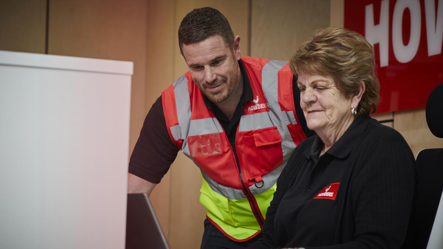 Two Howdens employees sat in front of a laptop. One is wearing a branded jacket and the other is in a high-vis vest.