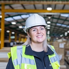 Howdens engineering staff in a large warehouse, wearing a hi-vis jacket and white hard hat.