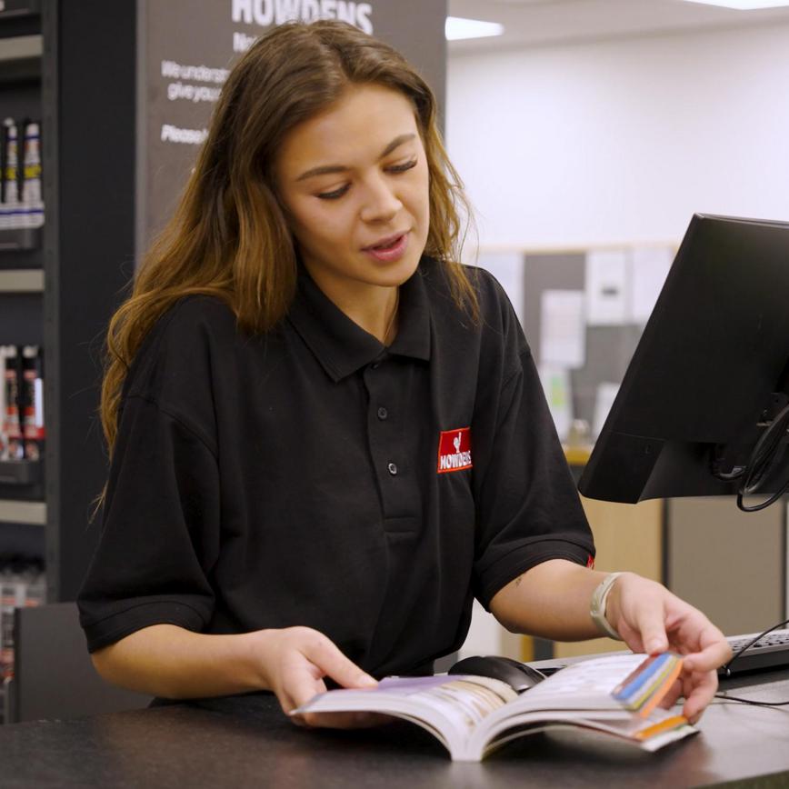 A Howdens apprentice depot staff member working as a counter sales employee. They are showing a customer a Howdens brochure.