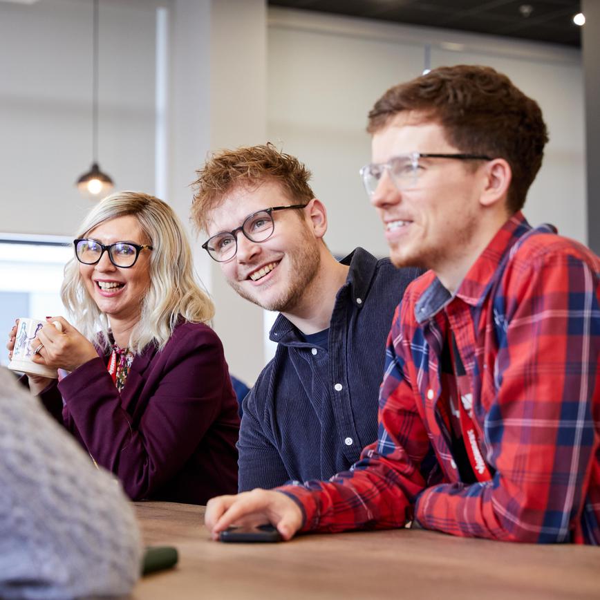 Group of people sat at a desk, 2 men in the foreground with a woman in the background. All smiling