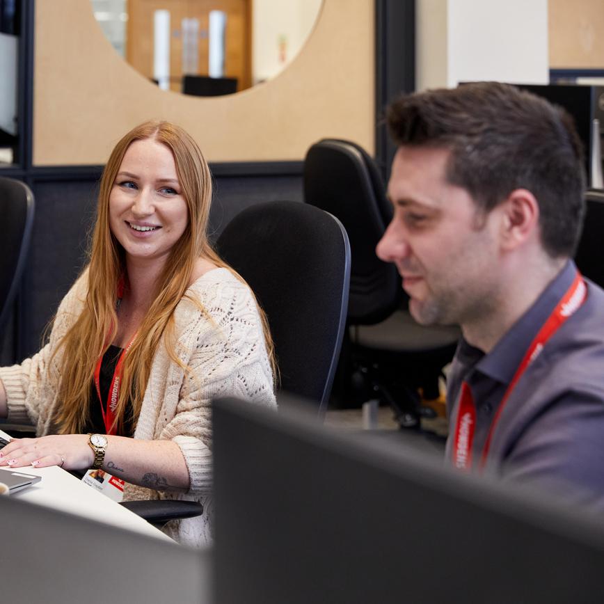 Man and woman sat at their computer desk, woman in the foreground and in focus smiling