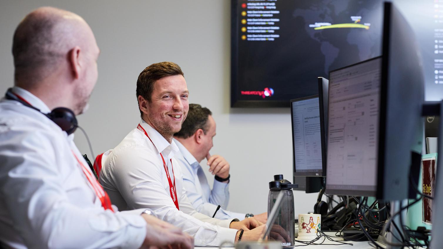 Three men sat at a computer desk, facing their monitors. The middle man is looking towards the camera smiling