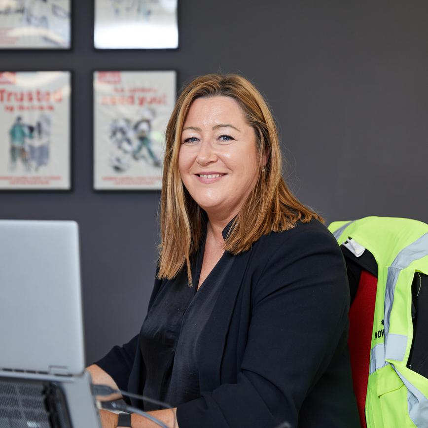 Woman sat at her desk with laptop in front of her, looking at the camera and smiling