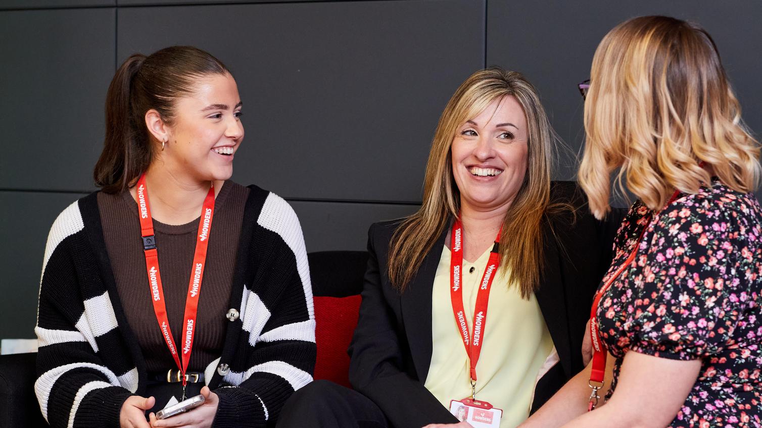 Three women sat down, looking at each-other and talking. The woman on the left can be seen smiling in focus