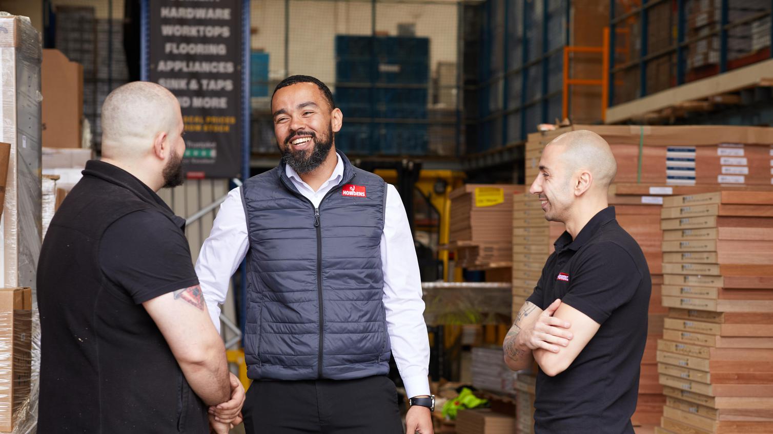 Three members of Howdens depot staff, stood talking in a warehouse with stock behind them.
