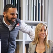Two Howdens staff members in an office setting, looking at a computer screen. Behind them are kitchen door colour samples.
